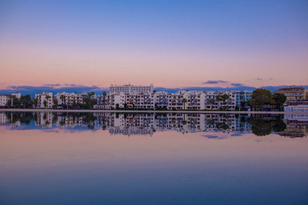 Una hermosa vista de la ciudad a través de la superficie del espejo del lago al amanecer. La ciudad de Port d 'Alcudia en Mallorca al amanecer. Hermoso gradiente rosa-azul en el cielo y el reflejo de la ciudad en el agua tranquila del lago. - Foto, Imagen