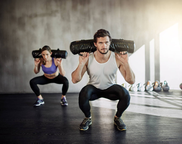 Άνδρας, γυναίκα και squat με sandbag για άσκηση γυμναστικής ή δύναμη ποδιών για την οικοδόμηση των μυών, πειθαρχία ή αθλητής. Προσωπικός γυμναστής, client and weightlifting class για αθλητικούς στόχους, προπόνηση ή γυμναστική. - Φωτογραφία, εικόνα