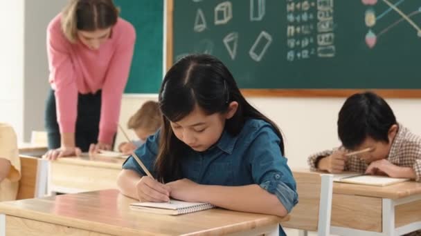 Ázsiai lány írja a tesztet, vagy órai munka közben a kamera az osztályteremben. Sokszínű boldog diák jegyzetel, és a tanár segít a kaukázusi fiú csinál tesztet, miközben ül a tábla előtt. Pedagógia. - Felvétel, videó