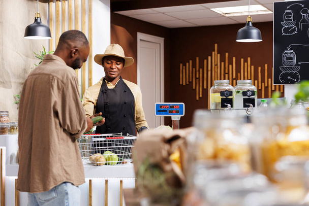 Öko-tudatos afro-amerikai vásárló érdeklődik az organikus, helyi eredetű gyümölcsökről és zöldségekről egy barátságos helyi szupermarketben. Egy kalapos eladó, aki az ügyfelét segíti a pénztárnál.. - Fotó, kép
