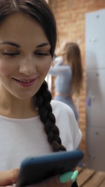 Vrouw gebruikt telefoon terwijl meisje vriend maakt kapsel staan in de buurt spiegel in kleedkamer. Dames bereiden zich voor op intensieve fitnesstraining close-up Verticaal schot. - Video