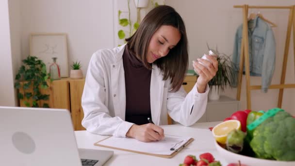 Mladá nutriční žena sedí za stolem s ovocem a zeleninou a pracuje na dietním plánu, který připravuje lékařský předpis pro pacienta v poradně. Zdravé stravování a výživa koncepce. - Záběry, video