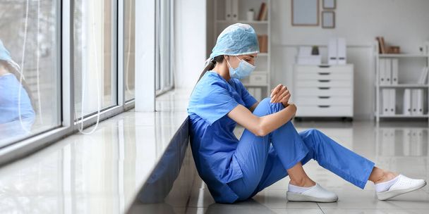 Stressed female surgeon sitting on floor near window in clinic - Foto, afbeelding