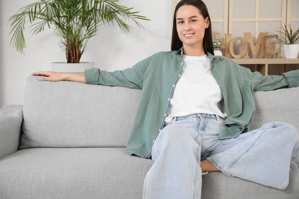 Hübsche junge Frau sitzt auf grauem Sofa im Wohnzimmer - Foto, Bild
