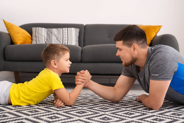 Young man with his little son having arm wrestling on floor at home - Fotó, kép