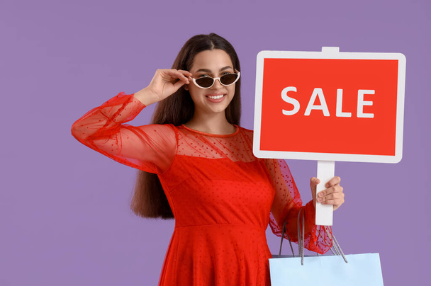 Beautiful young happy woman with shopping bags and SALE sign on purple background. Summer sale concept - Foto, imagen