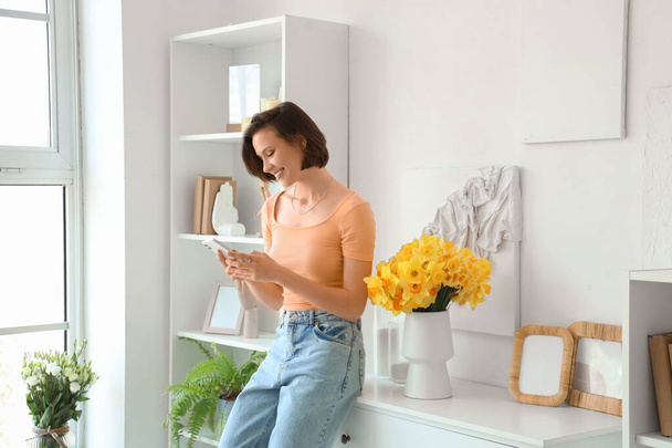 Young woman using mobile phone with daffodils in vase on commode at home - Foto, imagen