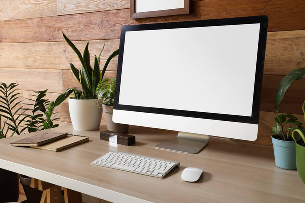 Lugar de trabajo con monitor de computadora en blanco y plantas verdes cerca de la pared de madera en la oficina - Foto, Imagen