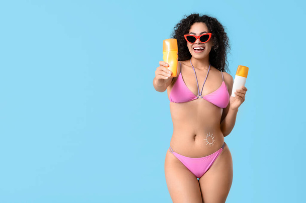 Beautiful young African-American woman in swimsuit with sun made of sunscreen cream on her belly against blue background - Photo, Image