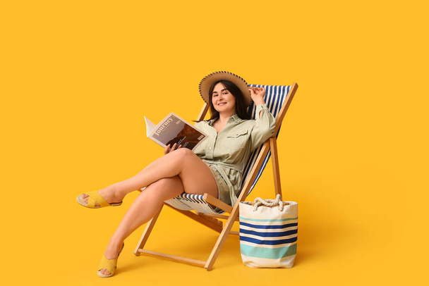 Beautiful young woman with magazine and stylish bag sitting in deck chair on yellow background - 写真・画像