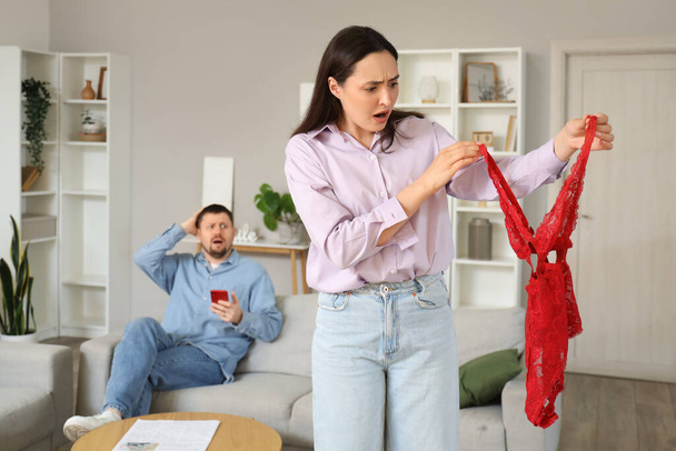Shocked young woman with found mistress's underwear and her husband at home. Cheating concept - Foto, imagen