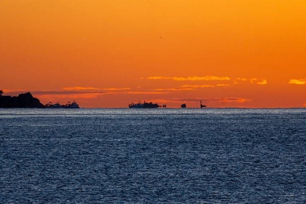 Silhouette di barche da pesca, in attesa di salpare, illuminate dal sole all'alba sulla linea dell'orizzonte del Mar Mediterraneo, creando miraggi e illusioni ottiche sotto un cielo arancione. - Foto, immagini