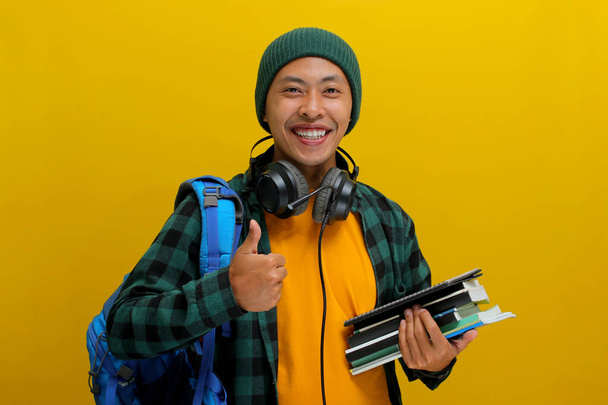 Estudante asiático confiante em gorro e roupas casuais, carregando mochila e fones de ouvido, Segurando uma pilha de livros. Isolado em fundo amarelo. Sucesso acadêmico, preparação ou a alegria de aprender. - Foto, Imagem