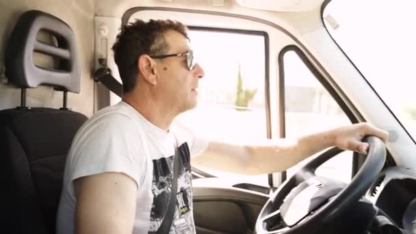 A man in sunglasses driving a truck in daylight turns the steering wheel with concentration. Driver in the process of driving a van in summer - Video, Çekim