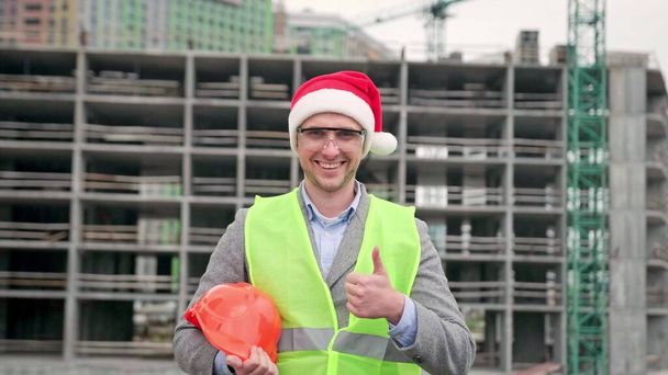 Smiling site supervisor in Santa hat holding protective helmet and giving thumbs-up while standing near unfinished multi-story house. Slow motion - Photo, Image