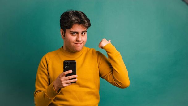 Portrait of happy Hispanic young male in yellow outfit holding smartphone while pointing at mockup space on green background - Photo, Image