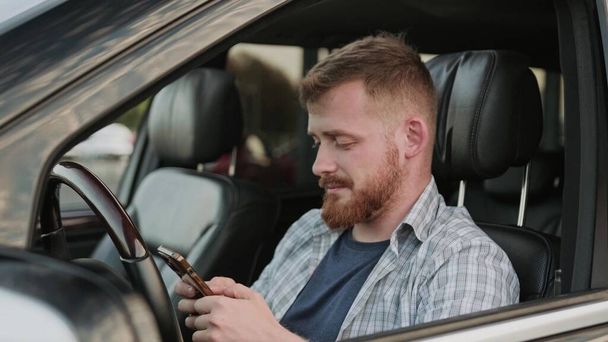 Ευτυχισμένος νεαρός που κάθεται στο αυτοκίνητο, ενώ πληκτρολογεί στο κινητό τηλέφωνο. Μεταφορές, τεχνολογία, ιδέα ταξιδιού. Αργή κίνηση - Φωτογραφία, εικόνα
