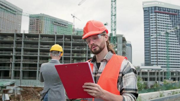 Cheerful foreman making notes on clipboard while posing for camera during site inspection with construction manager in background. Slow motion - Photo, Image