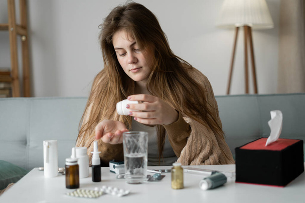 Caucasian young unhealthy woman sitting on a sofa in the living room, covered with a blanket, pours a pill from a bottle. Self-medication concept at home. - Foto, Bild