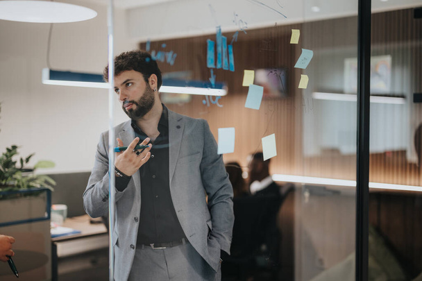 A focused businessman talks while gesturing, surrounded by sticky notes on a glass wall in a contemporary office, brainstorming ideas for sales and growth. - Photo, Image
