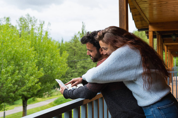 Vue latérale d'un couple de voyageurs enchantés embrassant sur la terrasse d'un chalet en bois et naviguant ensemble par téléphone portable pendant les vacances dans la chaîne de montagnes des Pyrénées - Photo, image