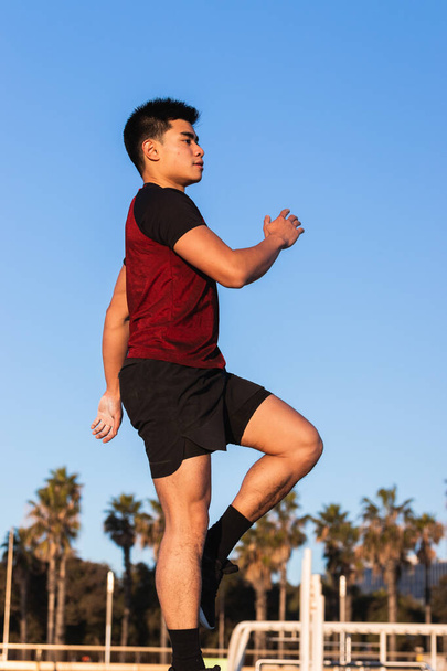 Vue latérale d'un jeune homme asiatique déterminé en vêtements de sport exécutant un pas au genou levez l'exercice sur un pneu pendant un entraînement de fitness extérieur - Photo, image