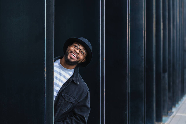 Optimistische volwassen zwarte hipster man met hoed en bril gluren uit de pilaar van de moderne stedelijke gebouw en kijken weg met nieuwsgierigheid - Foto, afbeelding