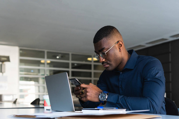 Boční pohled na Afroameričana soustředěný muž v brýlích a formální oblečení sedí v křesle u stolu při použití telefonu a netbooku v blízkosti dokumentu a odnést nápoj v lehkém pracovním prostoru - Fotografie, Obrázek