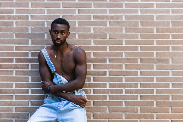 レンガの壁の背景とカメラを見ている街のデニムのオーバーオールに立っている筋肉の裸のトルソを持つアフリカ系アメリカ人男性 - 写真・画像