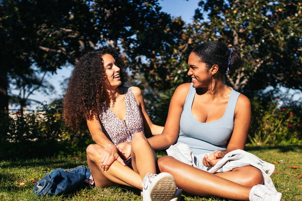 Kaksi afroamerikkalaista tyttöä istuu nurmikolla puistossa juttelemassa ja nauttimassa toistensa seurasta. He ovat lesbo pari elävät rakkautensa vapaasti. - Valokuva, kuva