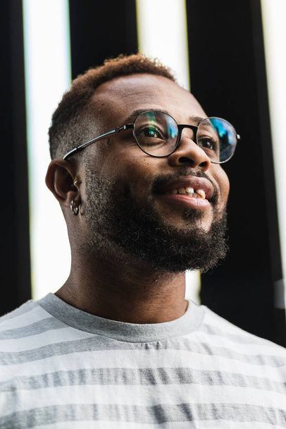 Alacsony szög pozitív magabiztos felnőtt afro-amerikai férfi szakáll és szemüveg visel alkalmi csíkos ing néz el a homályos háttér - Fotó, kép