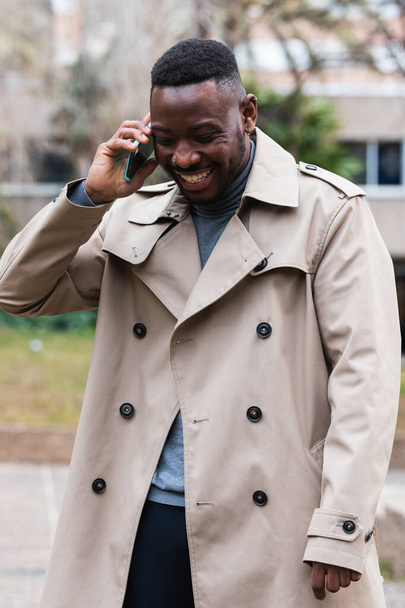 Hombre afroamericano adulto feliz con abrigo elegante hablando con el teléfono móvil en las manos mirando hacia otro lado mientras está parado en la calle urbana en el día de primavera - Foto, imagen