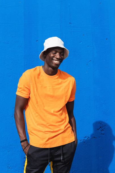 Nuori iloinen afroamerikkalainen mies kirkkaan keltainen t-paita ja valkoinen korkki poseeratessaan sinistä seinää vasten - Valokuva, kuva