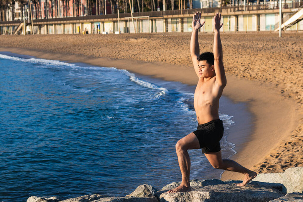 Tüm vücudu çıplak genç Asyalı bir erkeğin kumsalda yoga yaparken kollarını kaldırarak duruşu. - Fotoğraf, Görsel