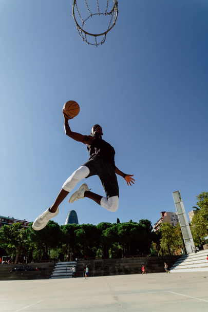 Low angle of fit zwarte mannelijke basketbalspeler in moment van springen boven de grond scoren bal in hoepel - Foto, afbeelding
