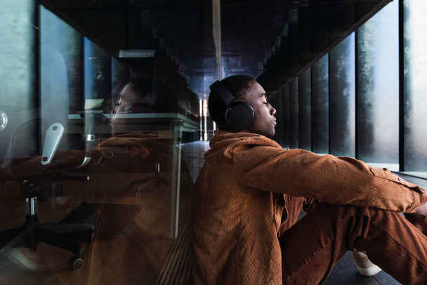 Vista lateral del hombre afroamericano escuchando música en auriculares inalámbricos sentados hacia atrás, una pared de vidrio con reflejo - Foto, Imagen