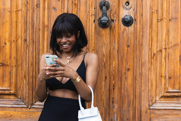 Усміхнена молода афроамериканка в чорному верхньому одязі з декольте повідомлення на смартфоні - Фото, зображення