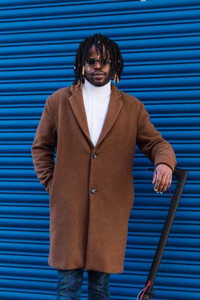 Homem hipster afro-americano adulto confiante com tranças afro vestindo casaco elegante e óculos de sol perto de scooter elétrico contra a parede listrada azul - Foto, Imagem