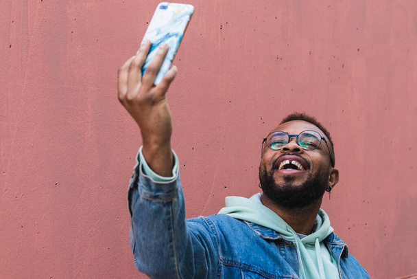 Низкий угол веселый взрослый черный хипстер парень в повседневной одежде делает автопортрет на мобильном телефоне на красном фоне - Фото, изображение