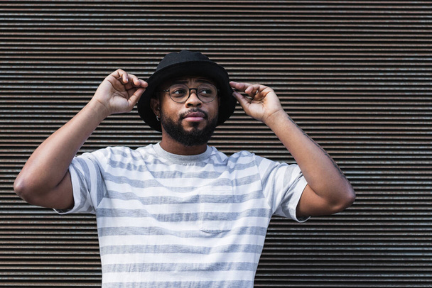 African American bearded guy with missing tooth in eyeglasses wearing trendy casual outfit and hat looking away while standing against shuttered wall - Photo, Image
