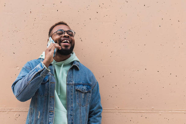 Взрослый хипстер бородатый афроамериканец в повседневной одежде и в очках, разговаривающий по телефону и смеющийся, довольный новостями, стоя напротив бежевой каменной стены на улице - Фото, изображение
