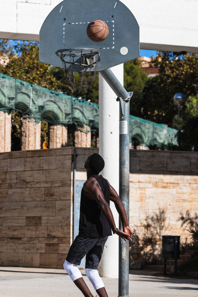 夏に裁判所でバスケットボールをするアフリカ系アメリカ人の男性アスリートのバックビューとカメラを見る - 写真・画像