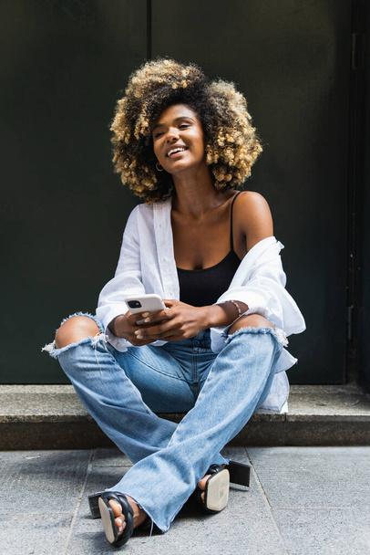 Volledig lichaam van positieve Afro-Amerikaanse vrouw in gescheurde jeans en wit shirt zitten met gekruiste benen en browsen mobiele telefoon - Foto, afbeelding