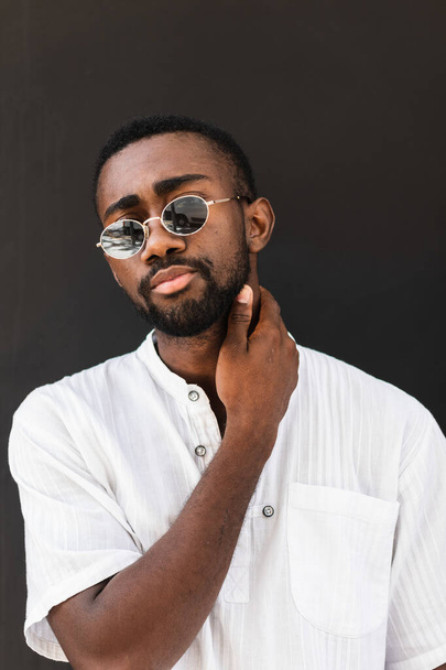 Confident African American male wearing stylish sunglasses and white t shirt standing on street while looking at camera and touching neck - Photo, Image