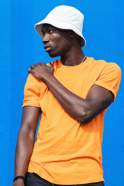Jonge Afro-Amerikaanse man in fel geel t-shirt en sportieve broek poseren terwijl staande tegen de blauwe muur - Foto, afbeelding