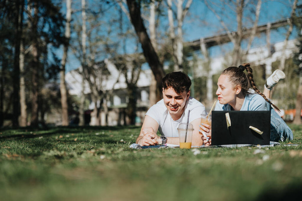 Uczuciowa para szkolna leżąca na trawie zaangażowana w naukę z książkami i laptopem, dzieląca chwilę wspólnoty. - Zdjęcie, obraz