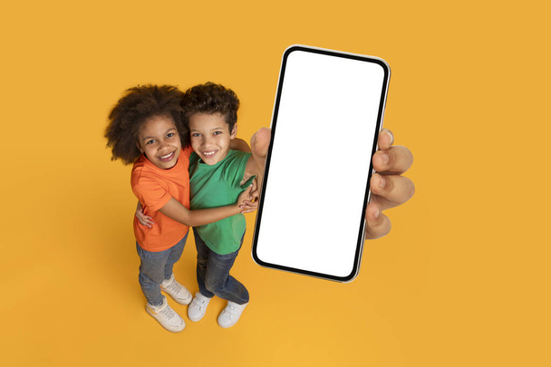 Dvě afroamerické děti, možná sourozenci nebo přátelé, stojí bok po boku a před sebou drží prázdnou obrazovku. Pozadí je zářivě žlutá barva, mockup, kopírovací prostor - Fotografie, Obrázek