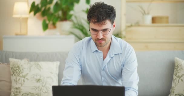 Uomo sorridente che digita su un computer portatile mentre siede comodamente sul divano di casa - Filmati, video