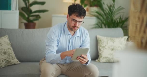 Homem trabalha em uma aplicação em um tablet digital enquanto sentado confortavelmente no sofá - Filmagem, Vídeo
