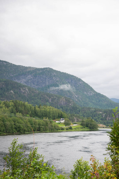 川岸の青空の下に緑豊かな木々が並ぶノルウェーの山々を望む美しい風景. - 写真・画像
